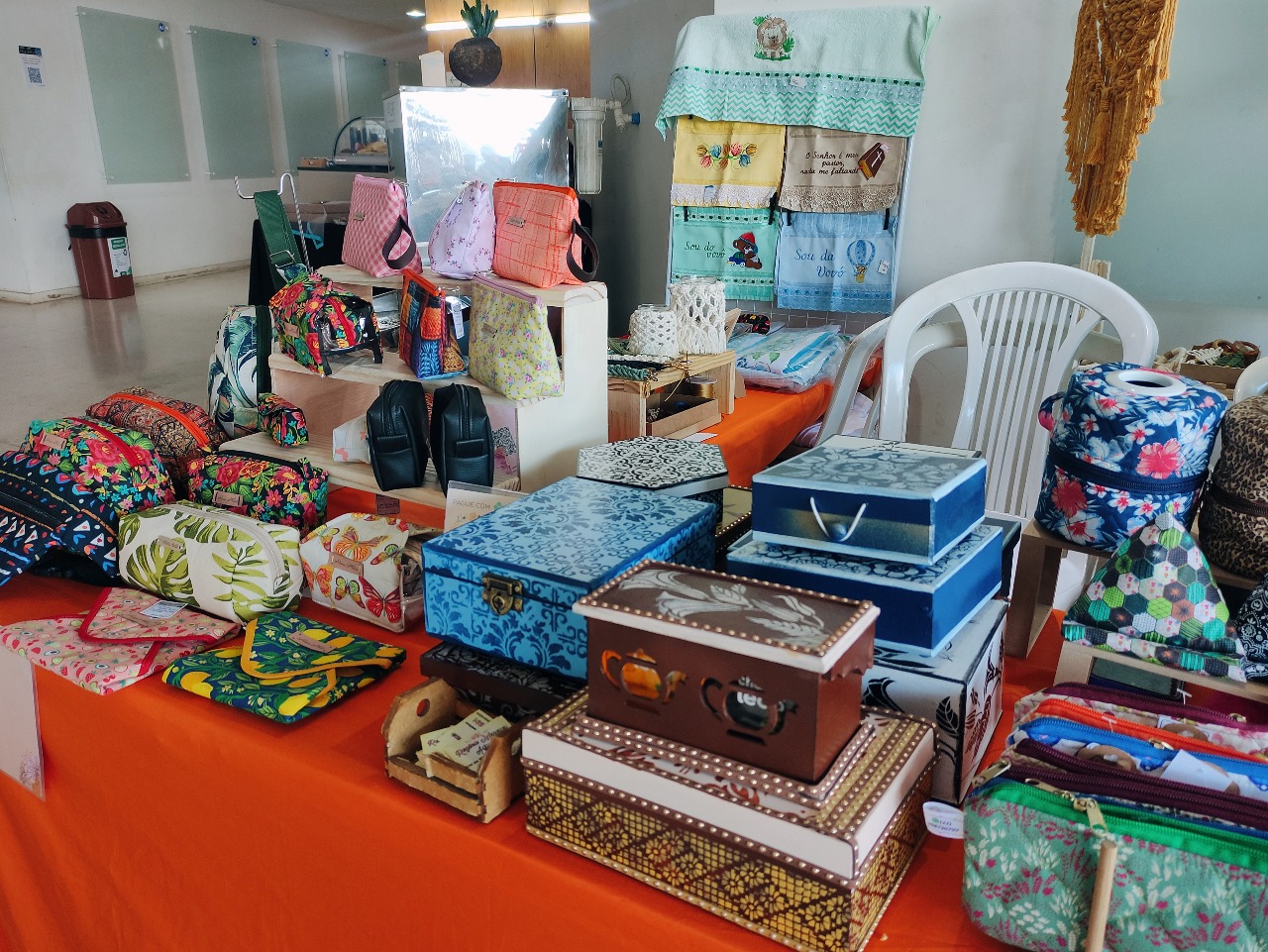 Mesa expositora cheia de necessaires e porta‑objetos de tecido estampado
(flores, folhagens e xadrez), caixas decorativas em madeira estampada nos tons azul e
marrom, além de panos de prato bordados pendurados num varal ao fundo