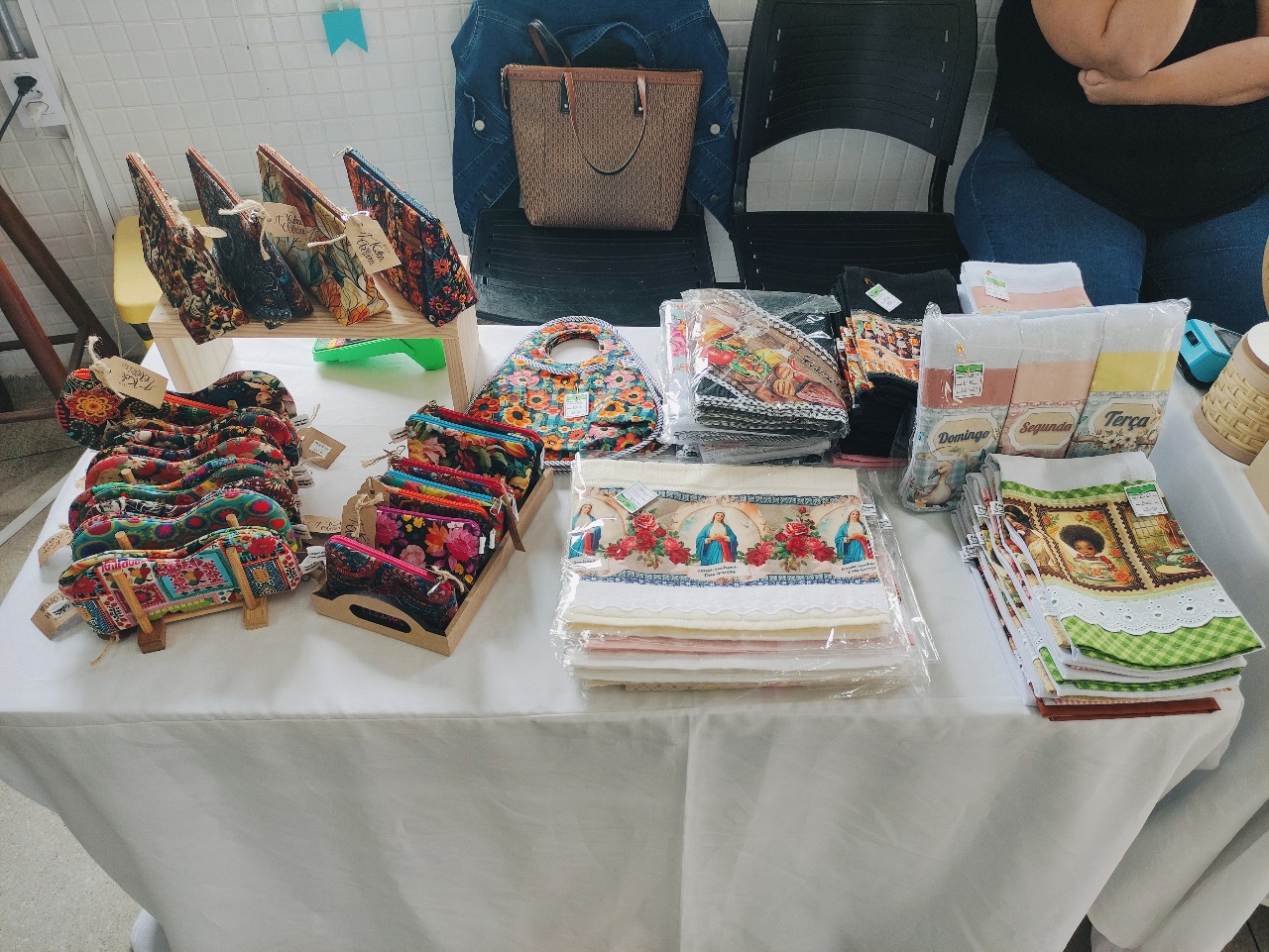 Mesa expositora com porta‑moedas e necessaires coloridas em tecidos estampados, xales e panos de prato com motivos religiosos, montados sobre toalha branca em salão de feira.
