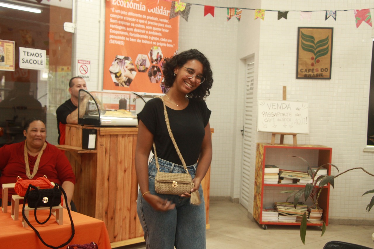 Mulher sorridente apresenta bolsas e artesanato expostos sobre mesa com toalha laranja, em frente a banqueta de madeira e painéis que exibem informações sobre economia solidária.