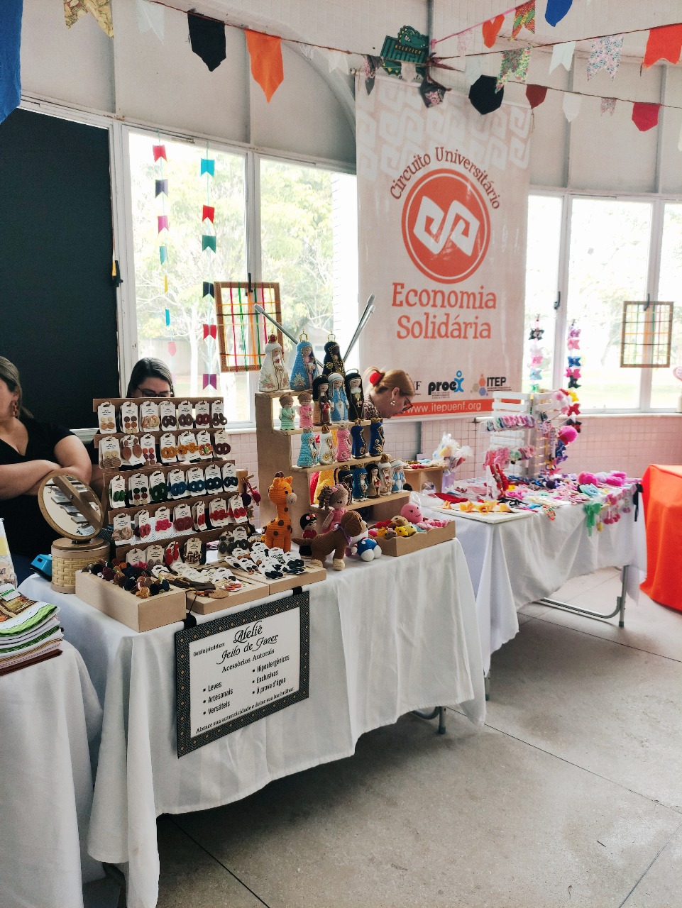 Barracas de feira cobertas por tecidos brancos em ambiente interno, com expositores mostrando bijuterias, bonecas de crochê e outros artesanatos, ao lado de banner vertical do ‘Circuito Universitário Economia Solidária’