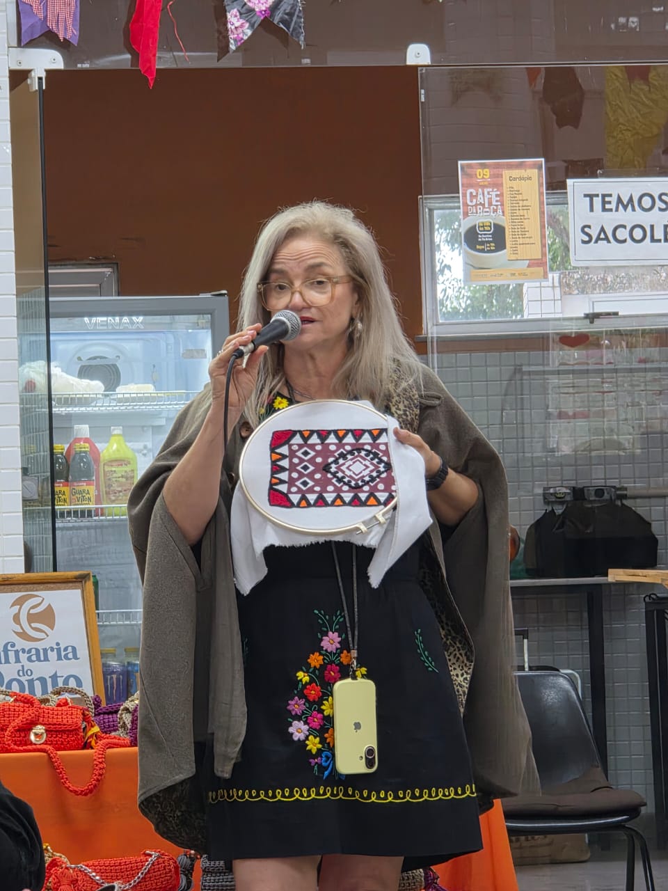 Mulher loira, de cabelos soltos e óculos, fala ao microfone segurando um bastidor de bordado com padrão geométrico vermelho, preto e branco. Atrás, mesa com toalha laranja exibe bolsas artesanais e um cartaz anunciando o evento.