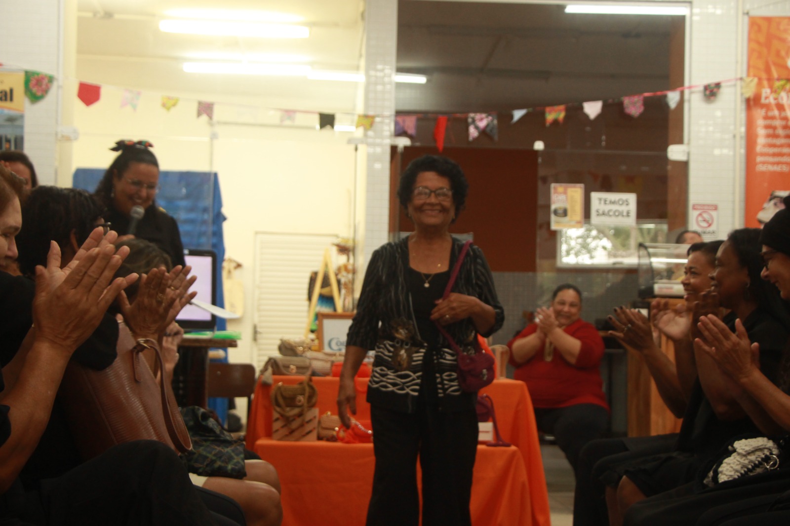 Mulher idosa sorri e caminha pelo centro de um salão decorado com bandeirolas de festa junina. Ela veste blusa preta com padrões brancos, calça preta e carrega uma pequena bolsa rosa-choque. À sua volta, pessoas sentadas em cadeiras de cada lado aplaudem entusiasmadas.
