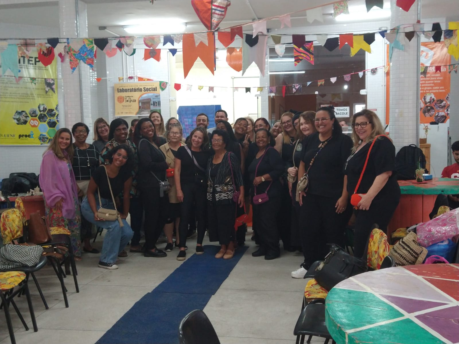 Foto de grupo com cerca de vinte participantes em um salão com decoração de bandeirolas coloridas. Todos posam lado a lado, muitos segurando bolsas de crochê; ao fundo, cartazes do Laboratório Social e ambiente arejado com mesas e cadeiras.