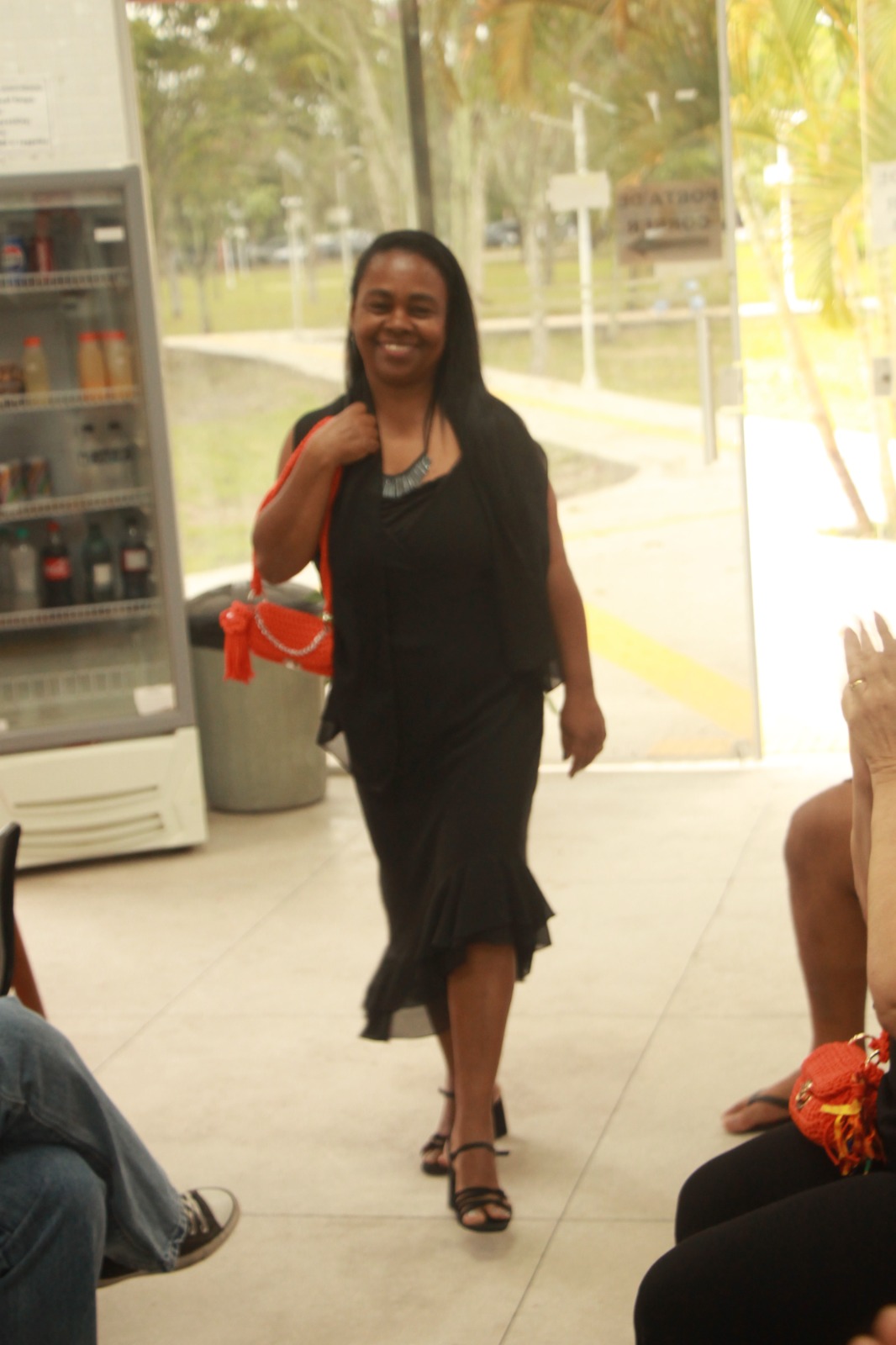 Mulher jovem, de vestido preto e bolsa de crochê laranja pendurada no ombro, avança pelo corredor de um ambiente interno, sorrindo em direção à câmera. Ao fundo, vê-se um bebedouro refrigerado e portas de vidro.