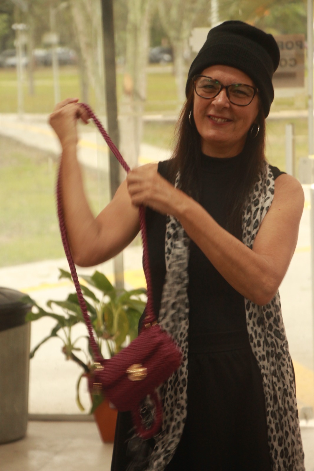 Mulher idosa sorri e caminha pelo centro de um salão decorado com bandeirolas de festa junina. Ela veste blusa preta com padrões brancos, calça preta e carrega uma pequena bolsa rosa-choque. À sua volta, pessoas sentadas em cadeiras de cada lado aplaudem entusiasmadas.