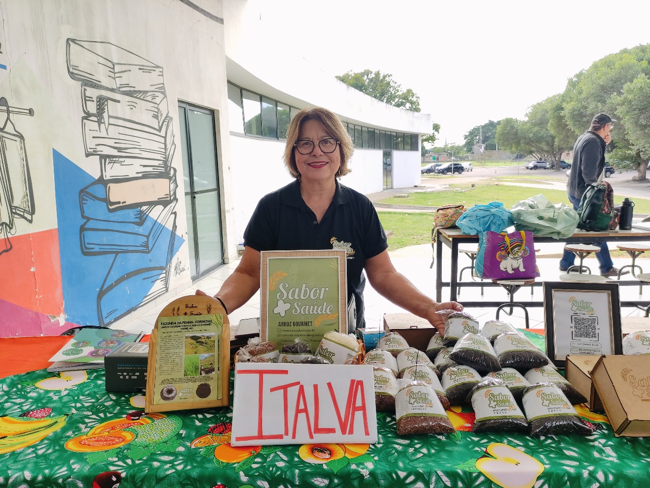 Fotografia de mulher sorridente, usando blusa preta, atrás de bancada exposta
      com pacotes de arroz gourmet “Sabor + Saúde” e placa com o nome “Italva”, sob um grafite
      de livros na parede da varanda do Restaurante Universitário.