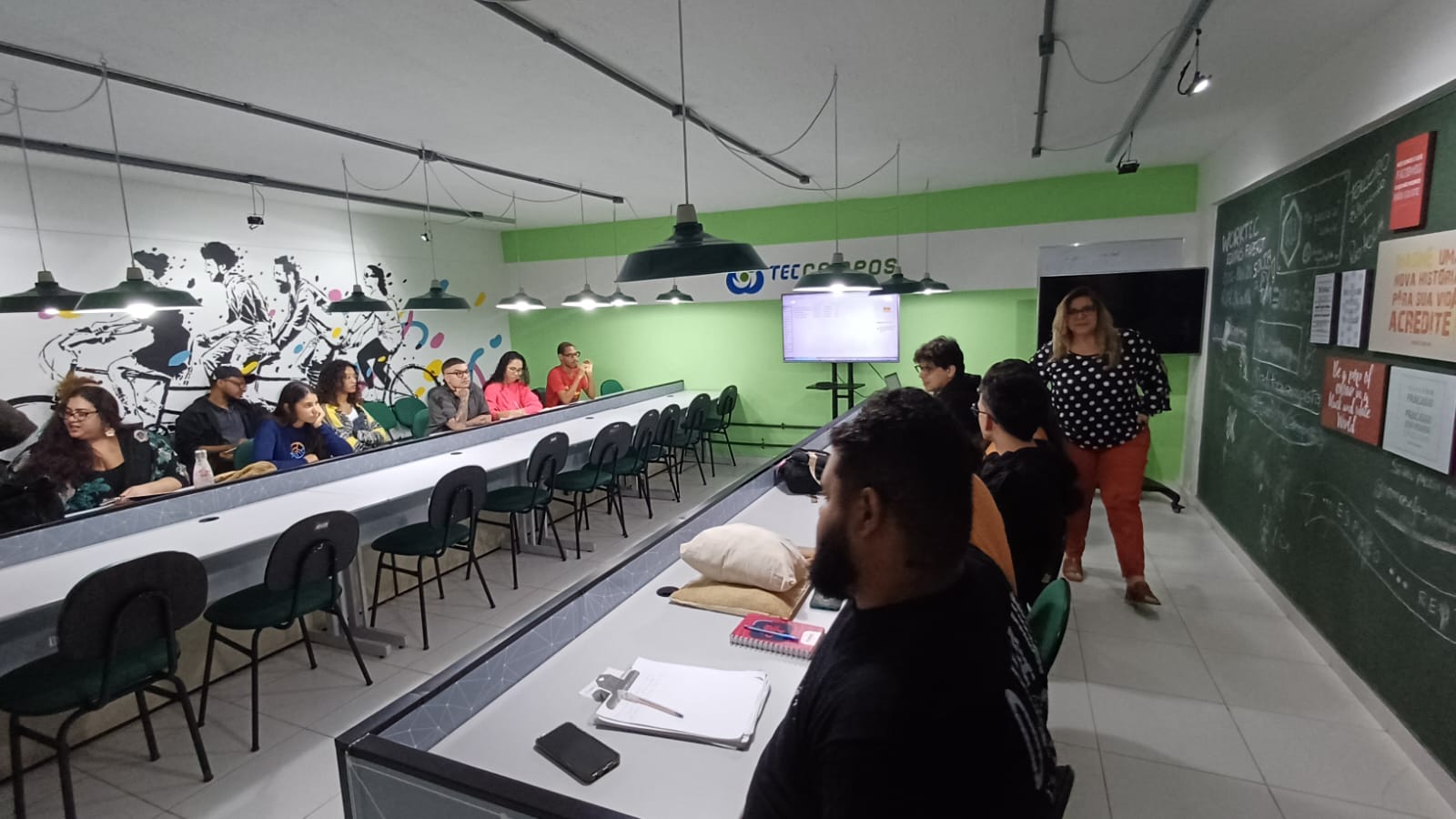 Alunos e alunas voltados para a frente, sentados em bancada em “U” de um laboratório criativo com parede verde e grafite. Instrutora em pé, próxima a lousa de cor escura repleta de anotações, conversa com o grupo.
