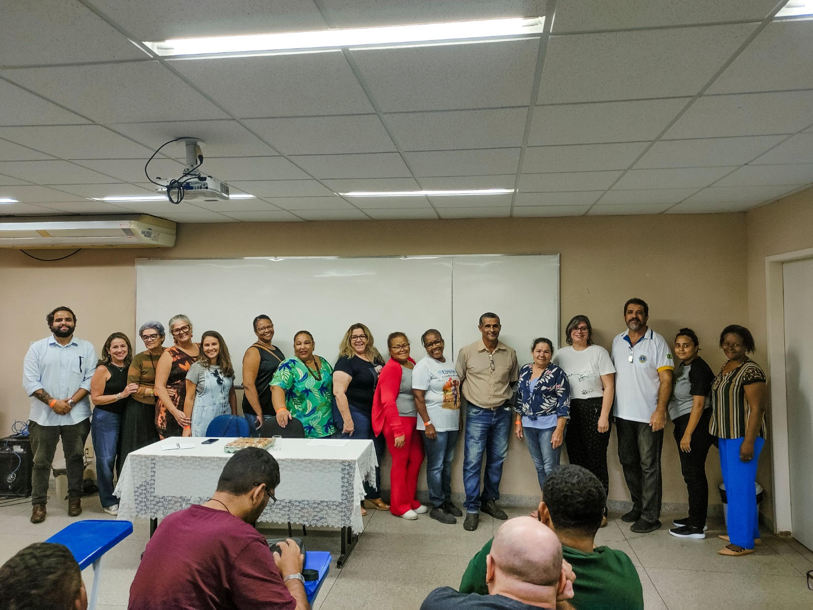 A imagem mostra seis pessoas sentadas ao redor de uma mesa oval em uma sala de reuniões. O grupo, composto por produtores, representantes institucionais e apoiadores do evento, participa de um momento de organização e articulação para a Feira Regional de Produtores Agroecológicos. Sobre a mesa, documentos, cadernos, canetas e um notebook com participantes em videoconferência reforçam o ambiente de trabalho conjunto. O clima é descontraído, com sorrisos que evidenciam o engajamento coletivo e a força da articulação territorial