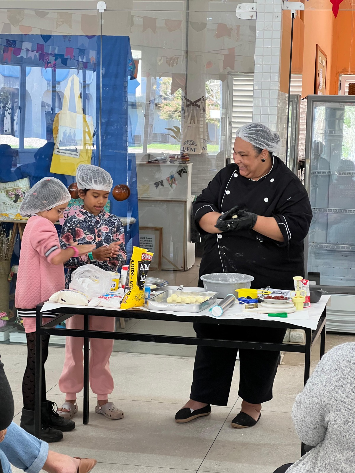 Demonstradora de uniforme preto e touca plástica sorri enquanto modela massa de biscoito em uma bancada. Duas crianças, também com toucas, ajudam a moldar bolinhas de massa, rodeadas por utensílios e ingredientes de confeitaria.