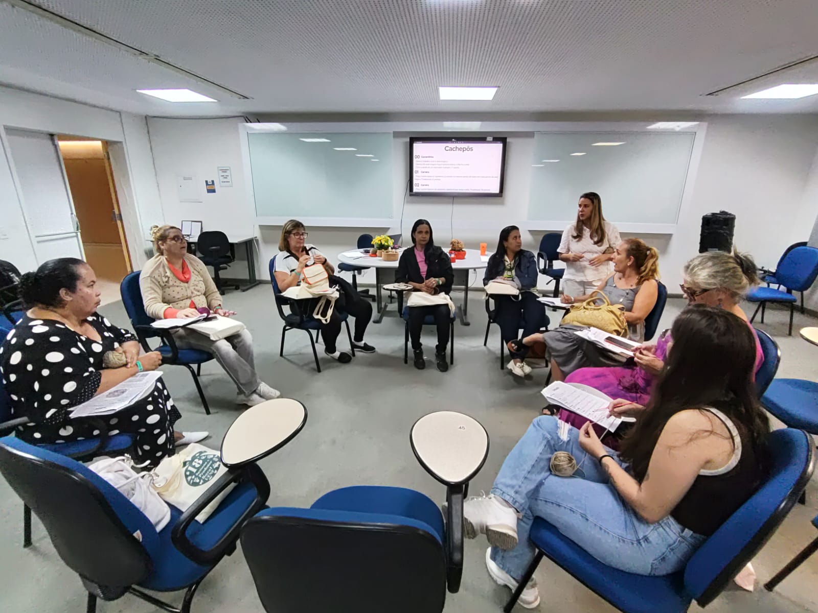 Sala de aula com as participantes sentadas em semicírculo em cadeiras giratórias azuis. No centro, uma instrutora fala, ao fundo mesa com cachepôs de crochê e televisão na parede exibindo o título “Cachepós”.