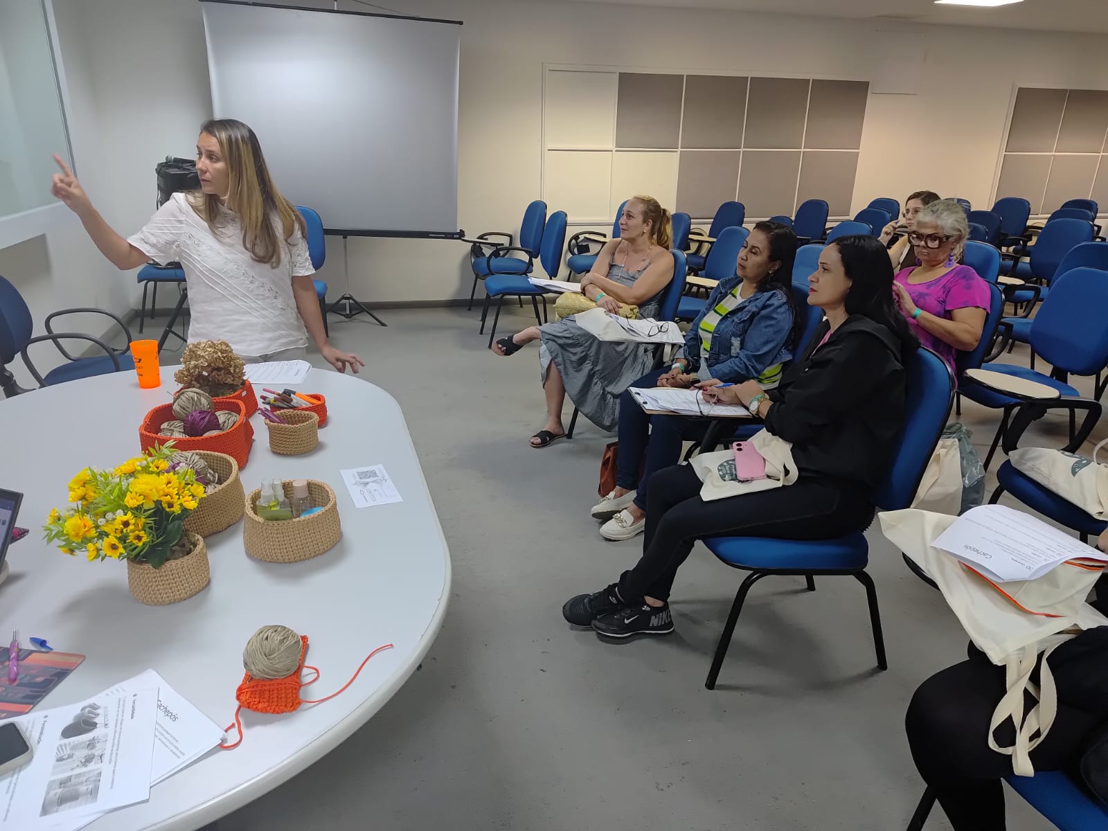 Mulher de pé, apontando para um quadro branco em sala de aula, dirige um pequeno grupo de alunas que está sentado em cadeiras azuis. À sua frente, sobre uma mesa oval, há diversos cachepôs de crochê em tons terrosos e coloridos, com novelos de fio e materiais de artesanato.