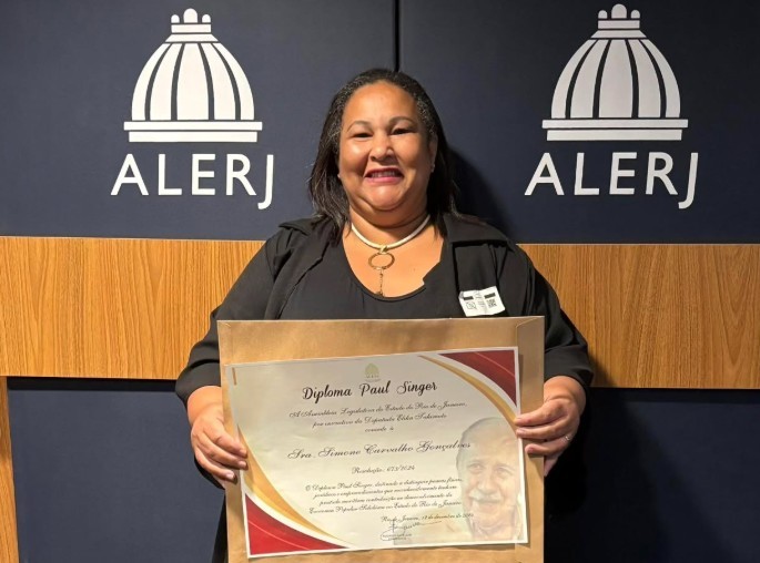 Simone Gonçalves sorri ao segurar o Diploma Paul Singer em frente a um painel com o logotipo da Alerj (Assembleia Legislativa do Estado do Rio de Janeiro). Ela veste roupa preta e está em pé, com expressão de orgulho e alegria, destacando a homenagem recebida por sua atuação na economia solidária.