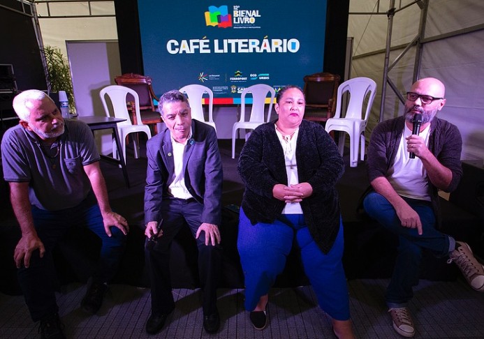 Durante a 12ª Bienal do Livro de Campos, Simone Gonçalves participa de uma mesa de debate no espaço Café Literário. Sentada ao centro, ela divide o palco com três homens, todos voltados para o público. O painel ao fundo exibe o nome do evento. O ambiente é informal e acolhedor, refletindo o tom da conversa sobre cultura, tradição e economia solidária.