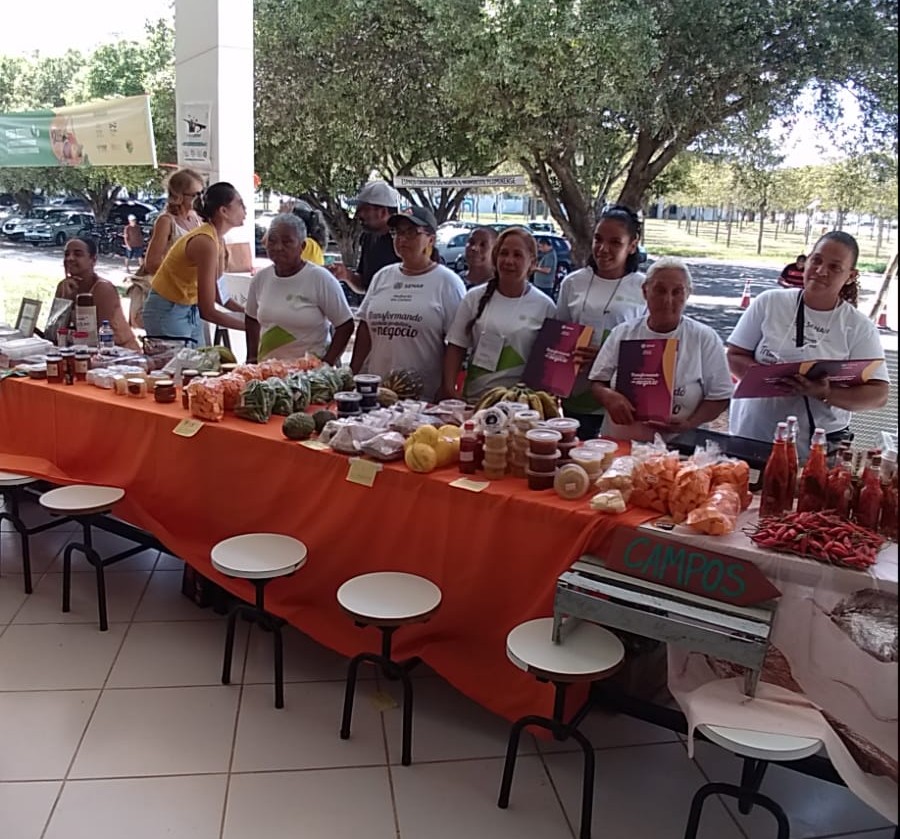 Na imagem, um grupo de mulheres e homens posa sorridente atrás de uma longa mesa forrada com tecido laranja. Sobre ela, uma variedade colorida de produtos agroecológicos — hortaliças, raízes, frutas, temperos, conservas, doces, cafés, garrafas com molhos e pimentas, além de peças de artesanato. Os expositores vestem camisetas brancas com logotipos institucionais e estão sob uma área coberta, com árvores ao fundo, transmitindo um clima de feira comunitária e colaborativa. À frente da mesa, um caixote com a inscrição “Campos” indica a origem dos produtos